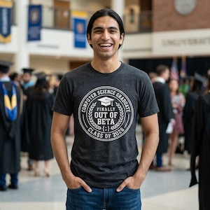 以下が含まれることがあります： 「COMPUTER SCIENCE GRADUATE FINALLY OUT OF BETA CLASS OF 2026」と書かれた白い円形グラフィックのダークグレーのTシャツ。 その人は笑顔で、青いジーンズを履いています。
