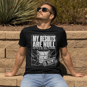 以下が含まれることがあります： 「MY RESULTS ARE NULL AND SO IS MY WILL TO DEBUG」というテキストと、ひび割れたデータベースのアイコン、そして「0 ROWS」と書かれた長方形のボックス、稲妻のエフェクトが描かれた白いグラフィックプリント付きの黒い半袖Tシャツ。