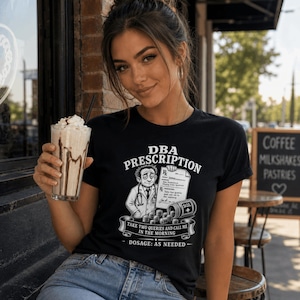Może przedstawiać: Czarny t-shirt z białą grafiką przedstawiającą lekarza trzymającego receptę z napisem DBA PRESCRIPTION. Projekt zawiera buteleczkę na tabletki z oznaczeniem SQL, stos tabletek oraz tekst TAKE TWO QUERIES AND CALL ME IN THE MORNING.