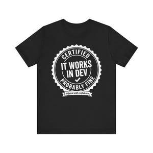 May include: Black t-shirt with a white circular graphic that reads "CERTIFIED IT WORKS IN DEV PROBABLY FINE" with a checkmark and the words "shipped with confidence" below. The t-shirt is made of a soft material.