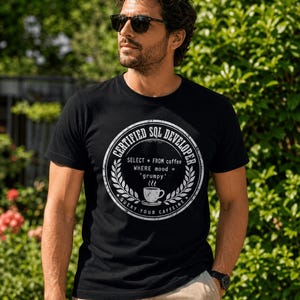 Puede incluir: Una camiseta negra de manga corta con un gráfico circular blanco que incluye el texto CERTIFIED SQL DEVELOPER, SELECT * FROM coffee WHERE mood = 'grumpy', y QUERY YOUR CAFFEINE, rodeando la ilustración de una taza de café humeante.