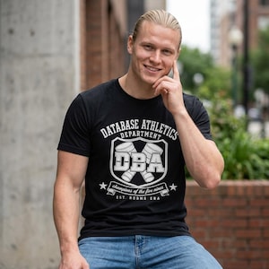 May include: Black crew neck t-shirt with a white graphic that reads "DATABASE ATHLETICS DEPARTMENT" above a shield design with "DBA" and two stylized barrels. Below the shield is a banner that reads "champions of the five nines" and "EST. RDBMS ERA."