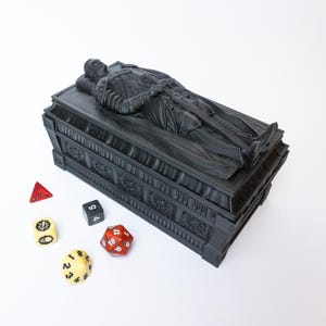 Puede incluir: Una caja rectangular negra con forma de sarcófago que presenta una figura esculpida de un caballero con armadura sobre la tapa. La caja tiene tallas laterales intrincadas y está acompañada por cinco dados de juego poliedros en rojo, negro y crema.