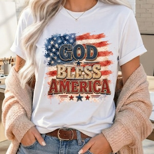 May include: White t-shirt featuring a distressed American flag design with the words "GOD BLESS AMERICA" in a patriotic font. The shirt has rolled-up sleeves and is paired with blue jeans and a brown belt.