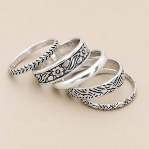 May include: A set of five silver-toned rings featuring various designs, including a thin band with a leaf pattern, a wider band with floral engravings, a smooth polished band, a band with textured diagonal lines, and a thin band with cross-hatching.