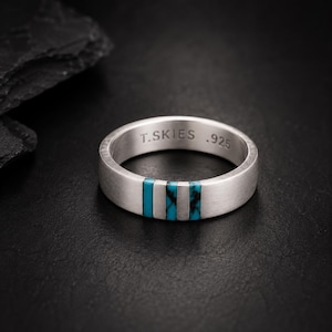 Op de afbeelding: Een zilveren bandring met een geborstelde afwerking en drie verticale inlays van blauw turkoois met zwarte matrix. De binnenzijde van de ring is gegraveerd met de tekst T.SKIES en .925, wat de samenstelling van sterling zilver aangeeft.
