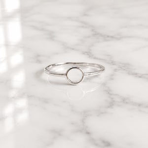 Op de afbeelding: Een zilveren ring met een minimalistisch design. De ring heeft een dunne band en een cirkelvormig open midden. De ring wordt gepresenteerd op een wit marmeren oppervlak, dat licht reflecteert. Dit sieraad is geschikt voor dagelijks gebruik.