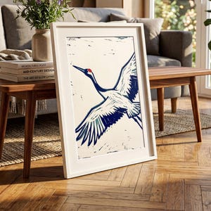May include: A framed art print featuring a crane in flight, rendered in blue and white. The bird has a red head and is set against a cream background. The print is displayed on a wooden table in a living room setting.