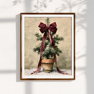 May include: A framed wall art print featuring a small evergreen tree in a terracotta pot. The tree is adorned with a large, deep burgundy velvet ribbon tied in a bow at the top, with long tails draping down the sides of the pot.