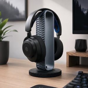 May include: A black and gray gaming headset rests on a gray and black headset stand. The stand has a textured design and a circular base. The headset has black earcups and a gray headband with blue accents. The SteelSeries logo is visible.
