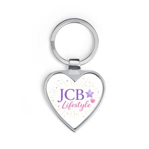 Puede incluir: Llavero en forma de corazón con un anillo de metal plateado. El corazón blanco presenta el texto "JCB Lifestyle" en morado y rosa, con una estrella morada y un corazón rosa, rodeado de rayos dorados.