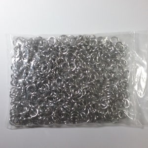 Op de afbeelding: Een doorzichtige plastic zak gevuld met een grote hoeveelheid zilveren springringen. De ringen zijn klein en rond, en ze zijn allemaal even groot.