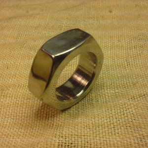 May include: A polished, silver-colored ring with a rectangular top and a rounded, octagonal shape. The ring is set against a textured, tan fabric background. The ring's design is simple and modern.