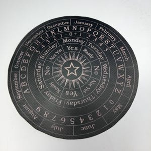Könnte beinhalten: Ein schwarzes, kreisförmiges Divinationsbrett mit einem Stern in der Mitte. Das Brett hat einen Ring mit Buchstaben, Zahlen, Wochentagen, Monaten des Jahres und den Wörtern "Ja", "Nein", "Bereit" und "Noch nicht".