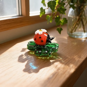May include: A glass figurine of a red ladybug with black spots and a white daisy flower on its back, resting on a translucent green four-leaf clover. The ladybug has a smooth, rounded body and the clover features detailed leaf segments.