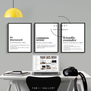 May include: Three black and white printable wall art prints with the definitions of the words "as discussed", "common sense", and "friendly reminder". The prints are framed and hanging on a white wall above a desk with a laptop, a camera, and a yellow lamp.