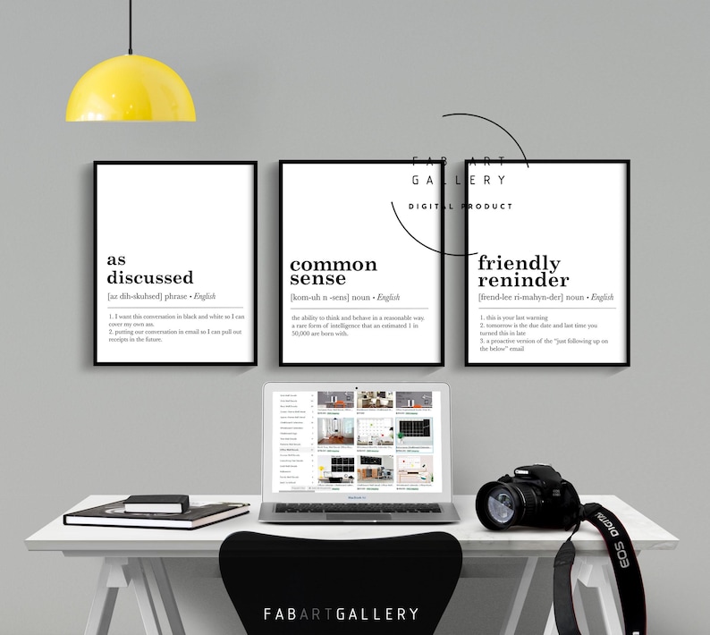 May include: Three black and white printable wall art prints with the definitions of the words "as discussed", "common sense", and "friendly reminder". The prints are framed and hanging on a white wall above a desk with a laptop, a camera, and a yellow lamp.