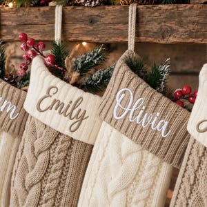 Op de afbeelding: Vier gepersonaliseerde kerstsokken hangen op een rij. Elke sok heeft een kabelgebreid patroon in afwisselende crème- en bruintinten met een geribbelde boord. De namen Liam, Emily, Olivia en Skylar zijn op de boorden geborduurd.