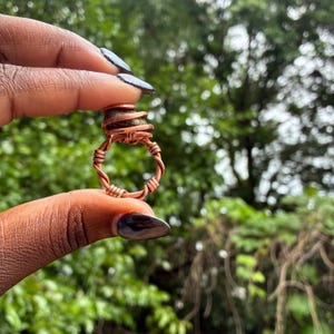 May include: A handcrafted copper wire ring with a natural stone centerpiece. The ring features a twisted wire band and a wrapped stone setting. The stone is a mix of green and brown hues. The ring is held up against a backdrop of green foliage.