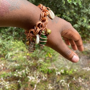 May include: A copper-colored chain bracelet with various charms. The charms include a small seashell, a green stone wrapped in copper wire, a small gold bell, and a glittery cylindrical charm. The bracelet is worn on a hand.