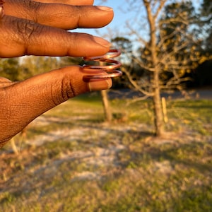 May include: A set of copper-colored spiral rings held between fingers. The rings are made of thin metal and have a coiled design. The background shows a blurred outdoor scene with trees and grass, lit by sunlight.