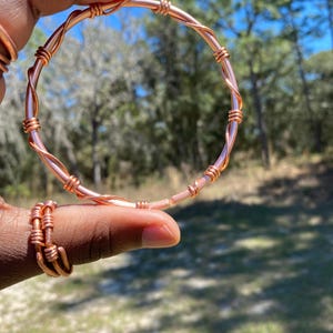 May include: Copper wire bracelet and ring set. The bracelet is a twisted design with a circular shape. The ring is also made of copper wire and has a similar twisted design. The jewelry is held up by a hand against a blurred background of trees and sky.