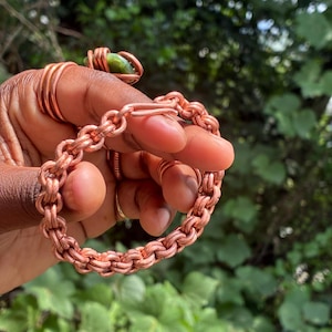 May include: A copper chain link bracelet featuring a hook and eye clasp mechanism. The bracelet is constructed from interlocking circular copper wire links, creating a textured, repeating pattern throughout the entire length of the jewelry piece.