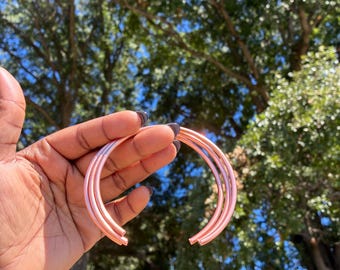 May include: A set of three smooth, rose gold-toned metal bangles with a circular, open-ended design. The bracelets feature a polished, reflective finish and a consistent, slender tubular construction that curves into a semi-circle shape.