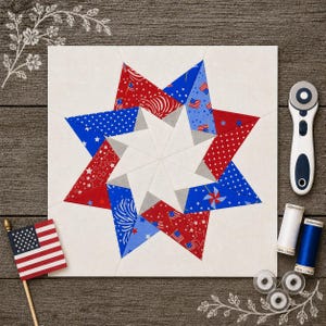 May include: A square, white fabric block with a starburst design in red, white, and blue patriotic patterns. An American flag, rotary cutter, and spools of thread are arranged around the block on a wood grain surface.