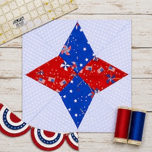 May include: A square quilt block with a star design in red and blue fabrics, featuring American flags and pinwheels. The block is surrounded by a light blue dotted fabric. Includes red and blue spools of thread and a quilting ruler.
