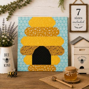 May include: A square quilt block pattern featuring a stylized yellow and gold beehive with a black entrance, set against a light blue background with a white circular pattern. The beehive design is composed of various yellow fabric textures.