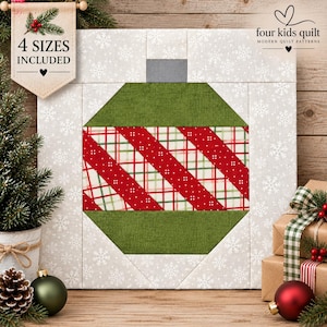 May include: A square quilt block featuring a central Christmas ornament design. The ornament has solid green top and bottom sections with a red, white, and green plaid middle band. The background is light grey with a white snowflake pattern.