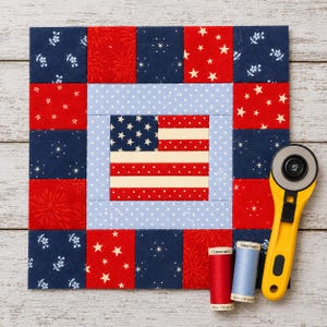 Pode incluir: Um bloco de colcha patriótico com a bandeira americana, com quadrados de tecido vermelho, branco e azul. Um cortador rotativo amarelo e bobinas de linha vermelha e azul estão ao lado da colcha. O bloco de colcha mede aproximadamente 25 cm quadrados.