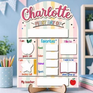 Könnte beinhalten: Ein gewölbtes Holzschild mit pastellfarbenen vertikalen Streifen und dem Namen Charlotte in rosa Schreibschrift. Es enthält leere Felder für den ersten Schultag, Alter, Größe, Lehrer und Favoriten wie Essen, Farbe, Tier und Sport sowie ein Buchsymbol.