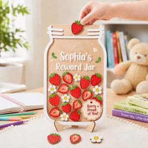 Peut inclure: Un bocal de récompense en bois en forme de pot Mason avec une bordure blanche, portant l'inscription 'Sophia's Reward Jar' et un panneau festonné indiquant 'Berry Proud of You!'. Il comprend plusieurs jetons fraises et fleurs.