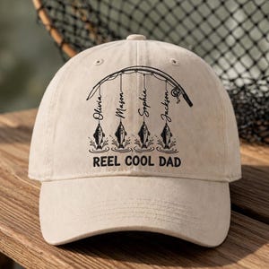 Op de afbeelding: Beige baseballpet met een gebogen klep. De pet heeft een hengelontwerp met de namen Olivia, Mason, Sophia en Jackson. De tekst "REEL COOL DAD" staat eronder.