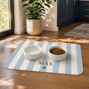Puede incluir: Un tapete de alimentación rectangular para mascotas con rayas verticales en azul claro y blanco. El tapete presenta el nombre MAX impreso en letras mayúsculas gris oscuro sobre un pequeño icono de huella gris oscuro. Dos cuencos blancos.