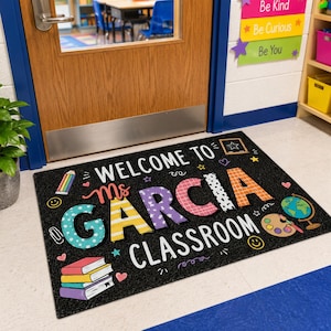 May include: A rectangular black classroom floor mat featuring the text "WELCOME TO Ms GARCIA CLASSROOM" in colorful, patterned letters. The design includes illustrations of a stack of books, a pencil, a globe, and a paint palette with brushes.