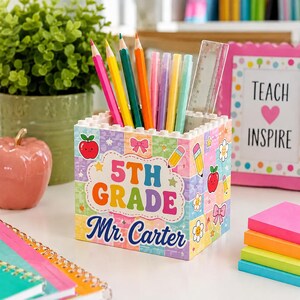 Könnte beinhalten: Ein quadratischer Stiftehalter aus Bausteinen mit einem bunten Karomuster, verziert mit Symbolen von Äpfeln, Stiften und Schleifen. Auf der Vorderseite steht der Text 5TH GRADE und Mr. Carter. Er enthält mehrere Buntstifte und ein durchsichtiges Lineal.