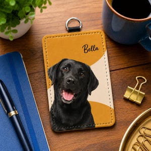 May include: A rectangular mustard yellow and cream ID badge holder featuring a portrait of a black Labrador retriever with its tongue out and the name Bella printed in black script text, topped with a silver metal D-ring attachment.
