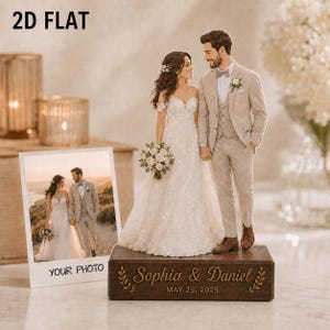 May include: A 2D flat wedding cake topper featuring a bride in a white lace gown and a groom in a beige suit standing on a rectangular wooden base. The base is engraved with the text "Sophia & Daniel" and "MAY 25, 2025" with leaf accents.