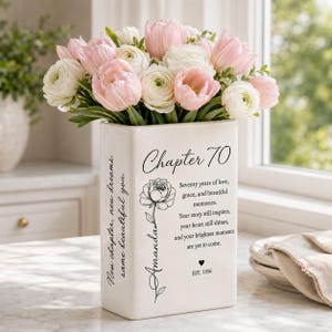 May include: A white rectangular ceramic vase filled with pink and white tulips and ranunculus. The front features black text reading Chapter 70, a floral illustration, the name Amanda, and a poem about seventy years of love and memories.