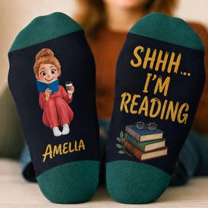 May include: A pair of dark navy blue socks with teal cuffs. The left sock features a cartoon illustration of a person reading a book while holding a coffee cup, with the name Amelia printed below. The right sock displays the text SHHH... I'M READING in yellow, with a stack of three books and a pair of glasses.