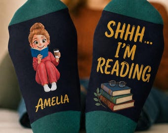 May include: A pair of dark navy blue socks with teal cuffs. The left sock features a cartoon illustration of a person reading a book while holding a coffee cup, with the name Amelia printed below. The right sock displays the text SHHH... I'M READING in yellow, with a stack of three books and a pair of glasses.