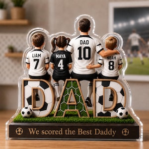 Puede incluir: Figura decorativa con temática de fútbol que representa a un padre y tres hijos con camisetas de fútbol. La palabra "DAD" se muestra de forma destacada con detalles de balón de fútbol. La base dice "We scored the Best Daddy."