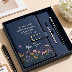 Op de afbeelding: Een marineblauwe notitieboekset in een bijpassende doos, met de tekst "She speaks with wisdom and teaches with kindness PROVERBS 31:26" en "Mrs. Taylor" met bloemmotieven. De set bevat twee zwarte pennen met zilverkleurige accenten.