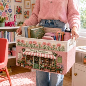 May include: A rectangular pink fabric storage bin with rope handles, featuring a printed illustration of a storefront labelled Olivia's Bookstore with a striped awning, flower boxes, and a top border of pink carnations filled with books.