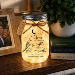 May include: A clear glass mason jar with a silver lid, filled with warm fairy lights. The jar features a black graphic of a cardinal on a branch, a crescent moon, stars, and the text: Gone from our sight, forever in our souls. Forever Remembered William. A brown twine bow and a small heart-shaped tag are tied around the neck.