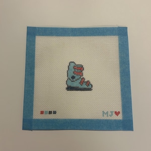 May include: A square needlepoint canvas featuring a light blue ice skate with red laces centered on a white grid. The canvas has a light blue fabric border. In the bottom left are small color squares, and in the bottom right is the text M.J. with a red heart.