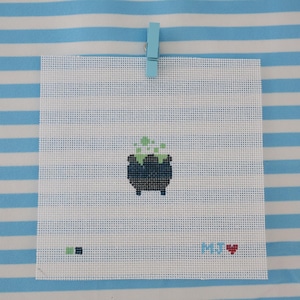 May include: A square piece of white needlepoint canvas featuring a cross-stitched dark blue cauldron with bubbling light green liquid. The canvas includes a small light blue clip at the top, color swatches, and the text MJ followed by a red heart.